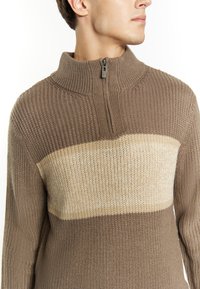 Khaki zip-hals trui met ribbelstructuur; heeft een crèmewitte horizontale streep over de borst. Vlak gebreide kraag met ritssluiting.