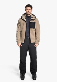 Veste en polaire beige avec une poche poitrine zippée noire, assortie à un pantalon noir. Convient pour les activités en extérieur, la texture semble douce.