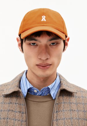 Orangenes Baumwollcap mit gebogenem Schirm und weißem gesticktem Logo. Getragen mit einer gemusterten braunen Jacke über einem blau gestreiften Hemd.