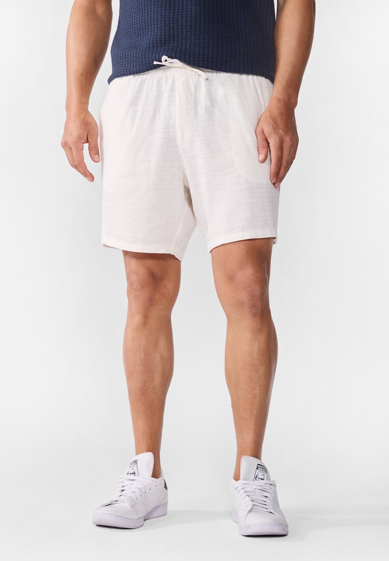 Shorts beige clair en tissu texturé avec une taille élastique et un cordon de serrage. Associés à des baskets blanches. Design simple et droit.