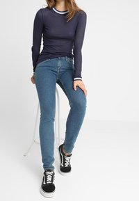 Marineblaues geripptes Langarmoberteil mit einem gestreiften Rundhalsausschnitt, kombiniert mit eng anliegenden blauen Jeans und schwarzen Sneakers mit weißen Akzenten.