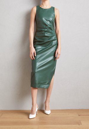 Femme portant une robe midi verte en cuir sans manches avec une fente latérale et des chaussures blanches à talons hauts, debout sur un sol en bois.