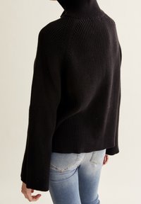 Svart ribbad polotröja med avspänt snitt och långa ärmar, designad med hög krage och kortad nederkant. Bärs med blå jeans.