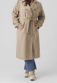 Beige trench-coat dengan pinggang berikat, kancing ganda, saku samping, dan epaulet. Dikenakan di atas jins denim biru model flare.