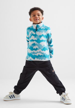 Child with curly hair wearing blue patterned fleece, black cargo pants, and white sneakers standing with legs apart on plain background.