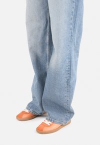 Zapatillas de ante naranja con cordones blancos y suelas de goma, combinadas con jeans holgados de lavado claro, que muestran un ajuste relajado y una textura suave.