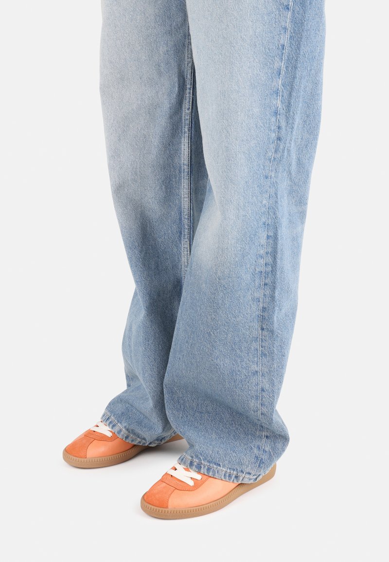 Zapatillas de ante naranja con cordones blancos y suelas de goma, combinadas con jeans holgados de lavado claro, que muestran un ajuste relajado y una textura suave.