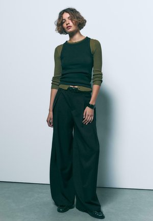 Femme aux cheveux courts et bouclés portant un haut à manches longues vert olive superposé à un débardeur noir, un pantalon large noir et des chaussures noires.