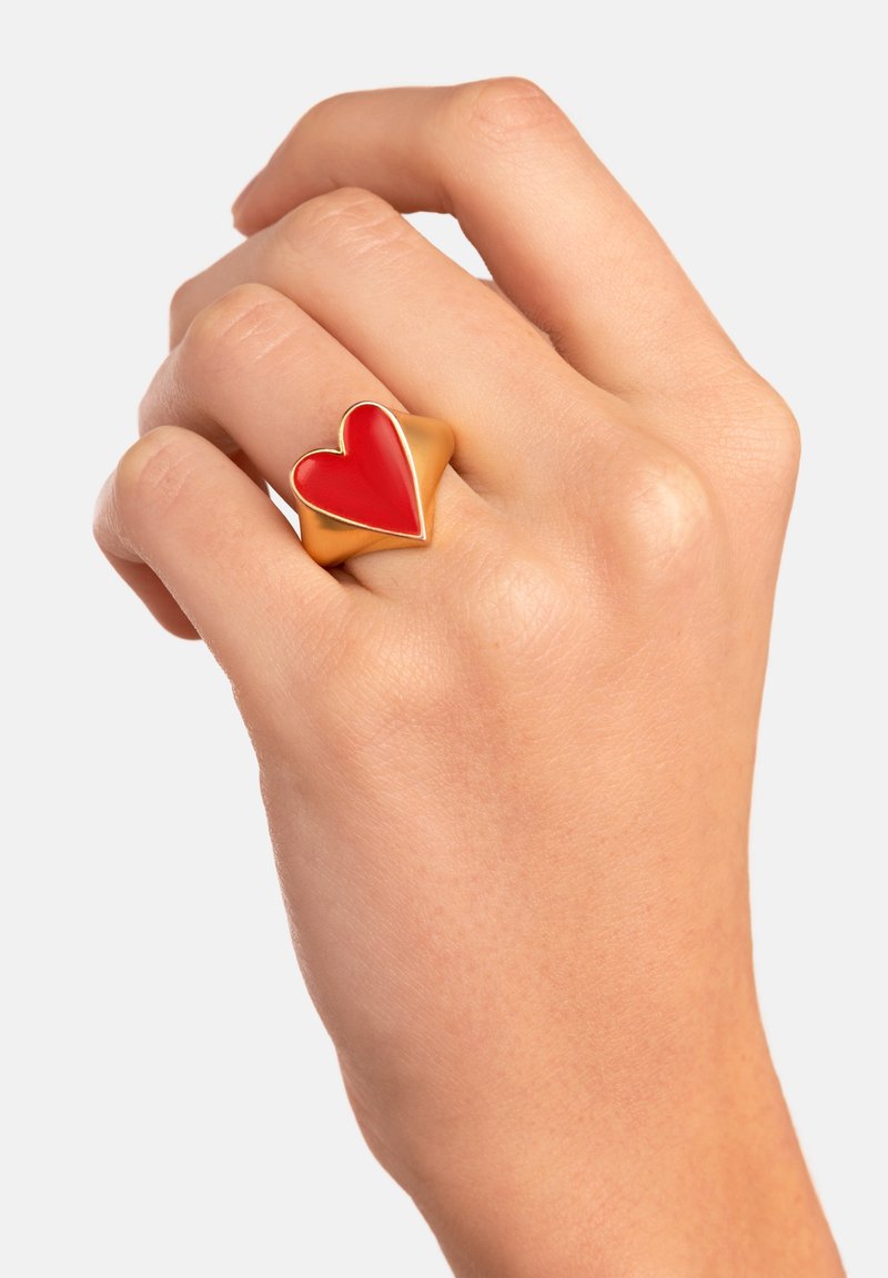 Main portant une bague en or avec un motif en forme de cœur rouge en relief sur le majeur sur fond blanc.