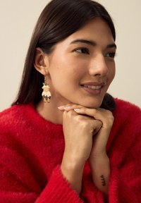 Kvinde i rød, fluffy sweater, hviler hagen på sammenfoldede hænder, iført festlige øreringe formet som pudding med detaljer af kristtorn, smilende let.