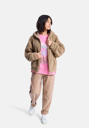 Veste zipée en peluche couleur beige avec une coupe décontractée, portée par-dessus un t-shirt graphique rose. Associée à un pantalon beige clair et des baskets blanches.