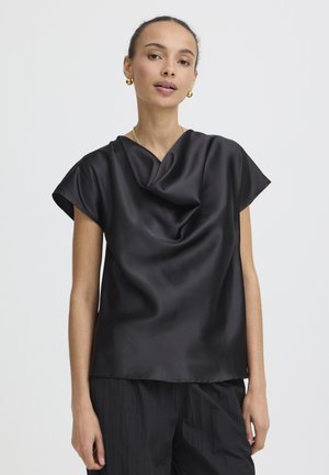 Femme portant un chemisier noir en satin à manches courtes et un pantalon noir ample, avec des boucles d'oreilles créoles dorées et un collier en or, fond neutre.