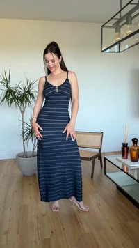 Femme en robe maxi sans manches bleu marine rayée posant à l'intérieur près d'une plante en pot, une chaise en bois et une table en verre avec des vases décoratifs.