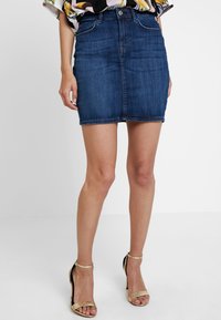 Jeanskjol i mörkblått med en figursydd midja, framfickor och sicksack-söm. Bärs med guldiga klacksandaler.