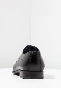 Chaussure habillée en cuir noir à finition lisse, bout arrondi, talon texturé et motif subtil sur la tige. Semelle basse et robuste.