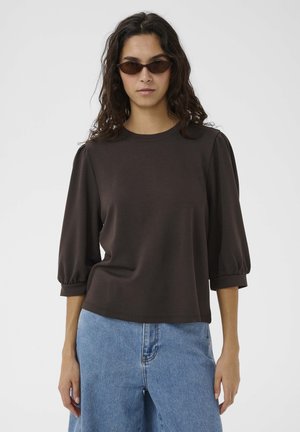 Donna con maglia marrone scuro con maniche a sbuffo, jeans azzurri e piccoli occhiali da sole marroni, in piedi davanti a uno sfondo bianco uniforme.