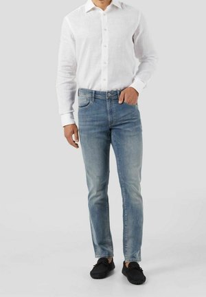 Mand iført en hvid langærmet skjorte med knapper, lyseblå slim-fit jeans og sorte slip-on sko, stående med den ene hånd i lommen.