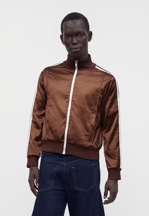 Mand iført en skinnende brun lynlåsjakke med hvide striber på ærmerne og mørkeblå jeans, stående foran en ensfarvet hvid baggrund.