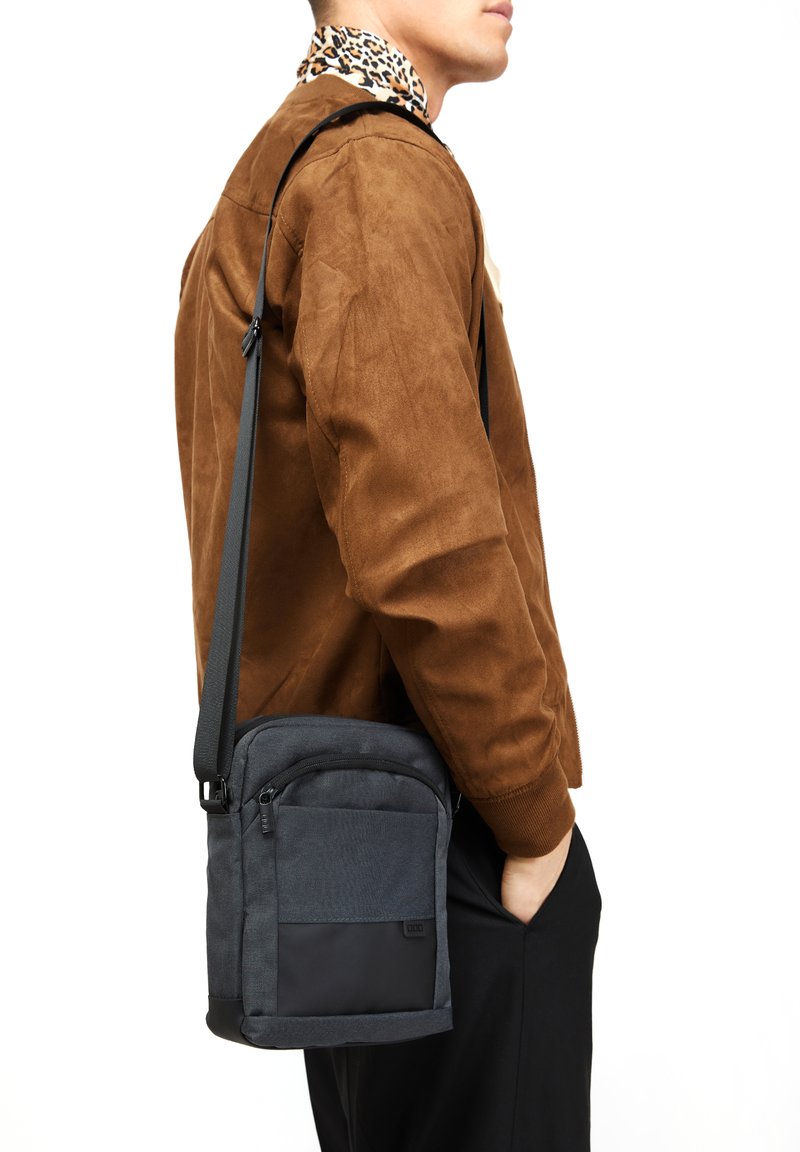 Tamnosivi crossbody torba s glatkom i teksturiranom tkaninom, s prednjim zip džepom i podesivim remenom, nosi se sa strane osobe.