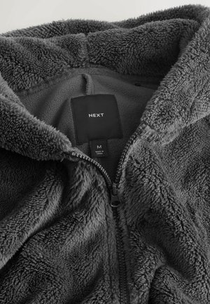 Grå fleece zip-up jacka med en mjuk, fluffig textur, med en etikett och dragkedjeslutning synlig på insidan. Storlek medium.