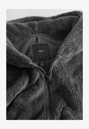 Szara polarowa kurtka z zamkiem błyskawicznym, o miękkiej, puszystej teksturze, z widoczną metką i zapięciem na suwak od wewnętrznej strony. Rozmiar średni.