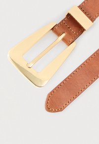 Καφέ δερμάτινη ζώνη με χρυσή τριγωνική αγκράφα και αντίστοιχο χρυσό κούμπωμα σε ανοιχτό φόντο.