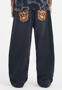 Black denim pants with a loose fit, featuring vivid embroidered skulls with fiery accents on the back pockets. Simple cuffed hem.