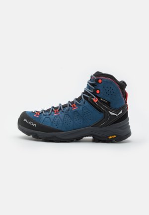 Scarpa da trekking blu con tomaia in suede, punta in gomma nera e suola Vibram. Presenta lacci grigi e dettagli arancioni. Design a metà altezza.