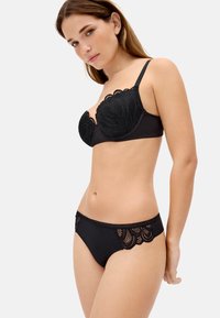 Mujer con conjunto de lencería de encaje negro, de pie y mirando ligeramente hacia la izquierda con expresión neutral sobre un fondo claro y liso.