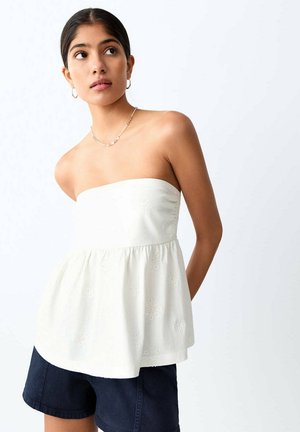 Mladá žena nosiaca kruhové náušnice, jemný retiazkový náhrdelník, biely top bez ramienok s kvetinovou výšivkou a tmavé šortky, pozerá sa do strany.