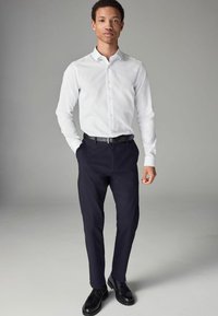 Cămașă albă cu nasturi, asortată cu pantaloni negri închis și pantofi negri. Cămașa are mâneci lungi cu manșete butonate și un guler clasic.