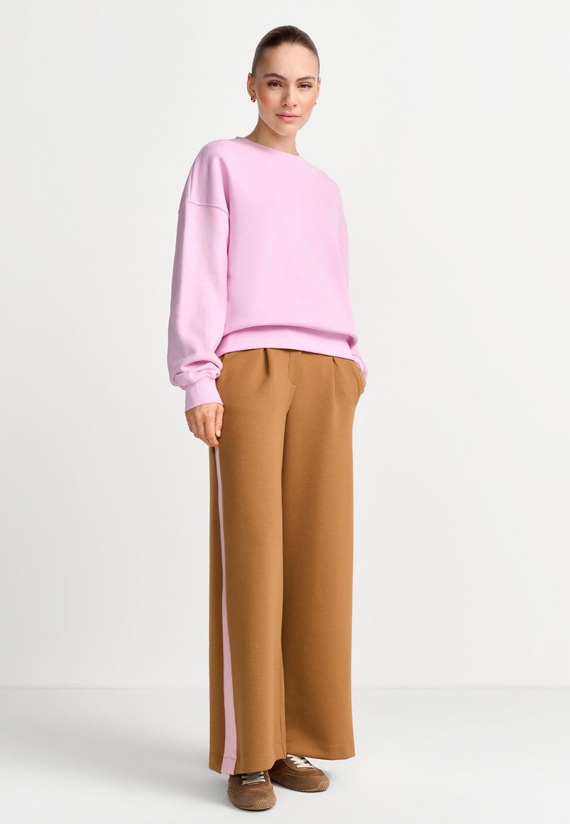 Žena v svetloružovej voľnej mikine, širokých hnedých nohaviciach s ružovými pruhmi po stranách a hnedých teniskách stojí pred jednoliatym bielym pozadím.