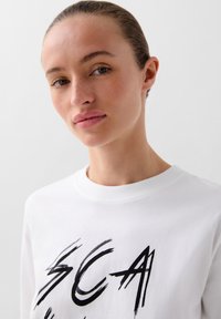 Hvid bomulds-sweatshirt med fed sort grafisk tekst. Ribbet rund hals og glat tekstur; nærbillede af en models overkrop.