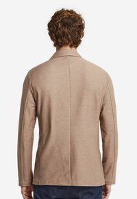 Beige skräddarsydd kavaj i mjukt tyg, slim fit, med en enda bakslits, långa ärmar, kavajkrage och sidofickor, bärs över blå jeans.