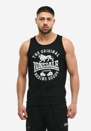 Hombre con camiseta sin mangas negra de Lonsdale London y pantalones cortos negros, de pie contra un fondo claro y liso.