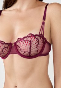Sujetador de encaje burdeos con copas transparentes que presentan bordados florales y bordes ondulados. Las correas ajustables y una banda de malla añaden detalle.