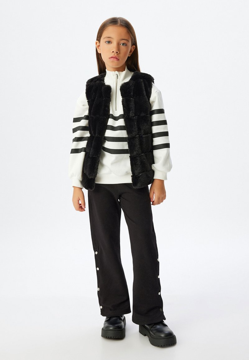 Gilet di pelliccia sintetica nera sopra un maglione bianco a righe nere, abbinato a pantaloni neri con bottoni a pressione. Scarpe nere chunky completano il look.