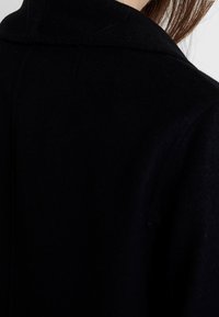 Černý vlněný kabát, zobrazuje detail zadního výstřihu a límce. Hladká textura s čistým designem bez viditelných kovových prvků nebo ozdob.