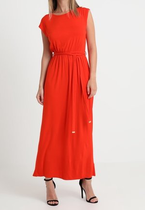 Femme portant une robe maxi rouge vif sans manches avec une ceinture nouée à la taille et des sandales à talons hauts noires, debout sur un fond blanc.