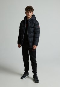 Veste matelassée noire avec capuche, fermeture éclair et écussons de logo. Associée à un pantalon de survêtement noir et des baskets grises. Design simple, matériau chaud.