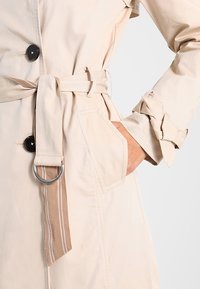 Beżowy płaszcz trencz z czarnymi guzikami, z paskiem w talii wykonanym z materiału w paski oraz ręką w bocznej kieszeni, rękawy z mankietami i paskami.