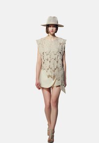 Top en dentelle beige avec un motif floral, superposé à un tunique crème. Assorti à un chapeau en paille léger et des sandales à lanières, arborant un design minimaliste.