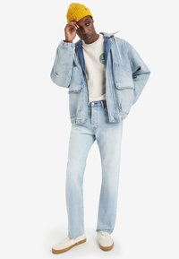 Light blue denim jacket with large pockets, paired with light blue jeans, white shirt, and yellow knitted beanie. White shoes with brown soles.