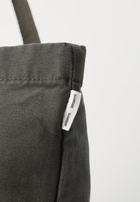 Mørkegrå totebag laget av teksturert stoff, med en liten hvit etikett med "SAMSOE SAMSOE" festet til sømmen på siden.