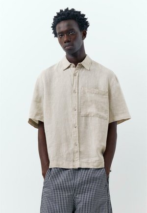 Tânăr purtând o cămașă bej cu mâneci scurte și buzunar și pantaloni bleumarin în carouri, stând cu mâinile în buzunare.