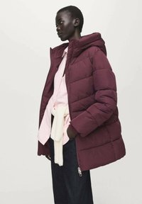 Vínově červená prošívaná péřová bunda s kapucí, s prošívaným vzorem a bočními zipovými kapsami, nošená přes růžovou košili a černé kalhoty.