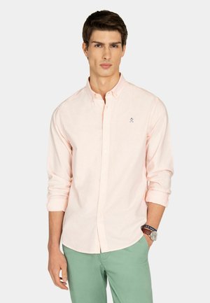 Joven con camisa de botones rosa claro y pantalones verdes, con una mano en el bolsillo y un reloj en la muñeca.