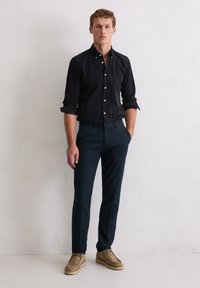 Schwarzes Hemd mit Knöpfen, dunkelblaue Hose, beiges Wildlederschuhe. Das Hemd hat eine schlanke Passform und hochgekrempelte Ärmel. Einfaches, sauberes Design.