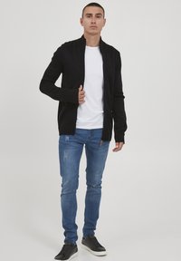 Svart hettejakke med glidelås, hvit T-skjorte under, blå slitte jeans og svarte joggesko. Strikket tekstur med ribbestrikkede mansjetter. Uformell passform.