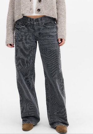 Femme portant un jean noir taille haute à jambes larges avec un motif texturé, associé à des chaussures marron et un cardigan beige court.